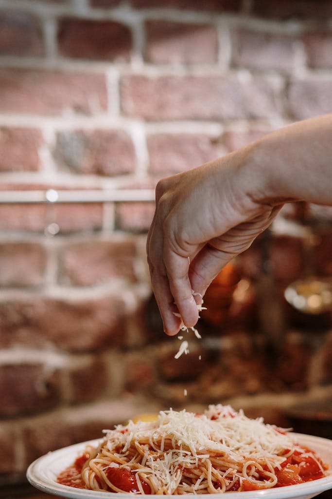 Close-up of a hand sprinkling cheese on spaghetti against a cozy brick background.